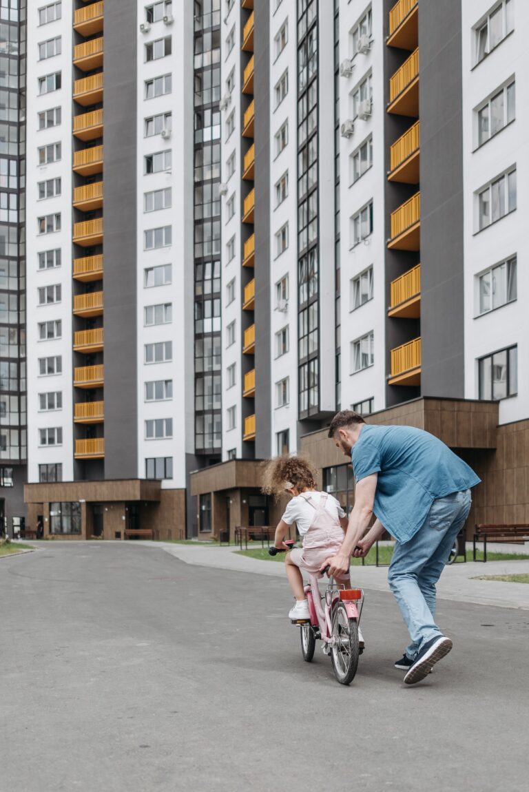 Father teaching daughter how to ride a bicycle in front of a block of flats.
