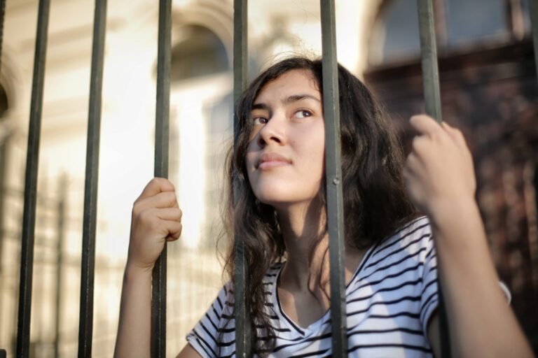 A person holding onto metal bars with their head against and looking through the bars.